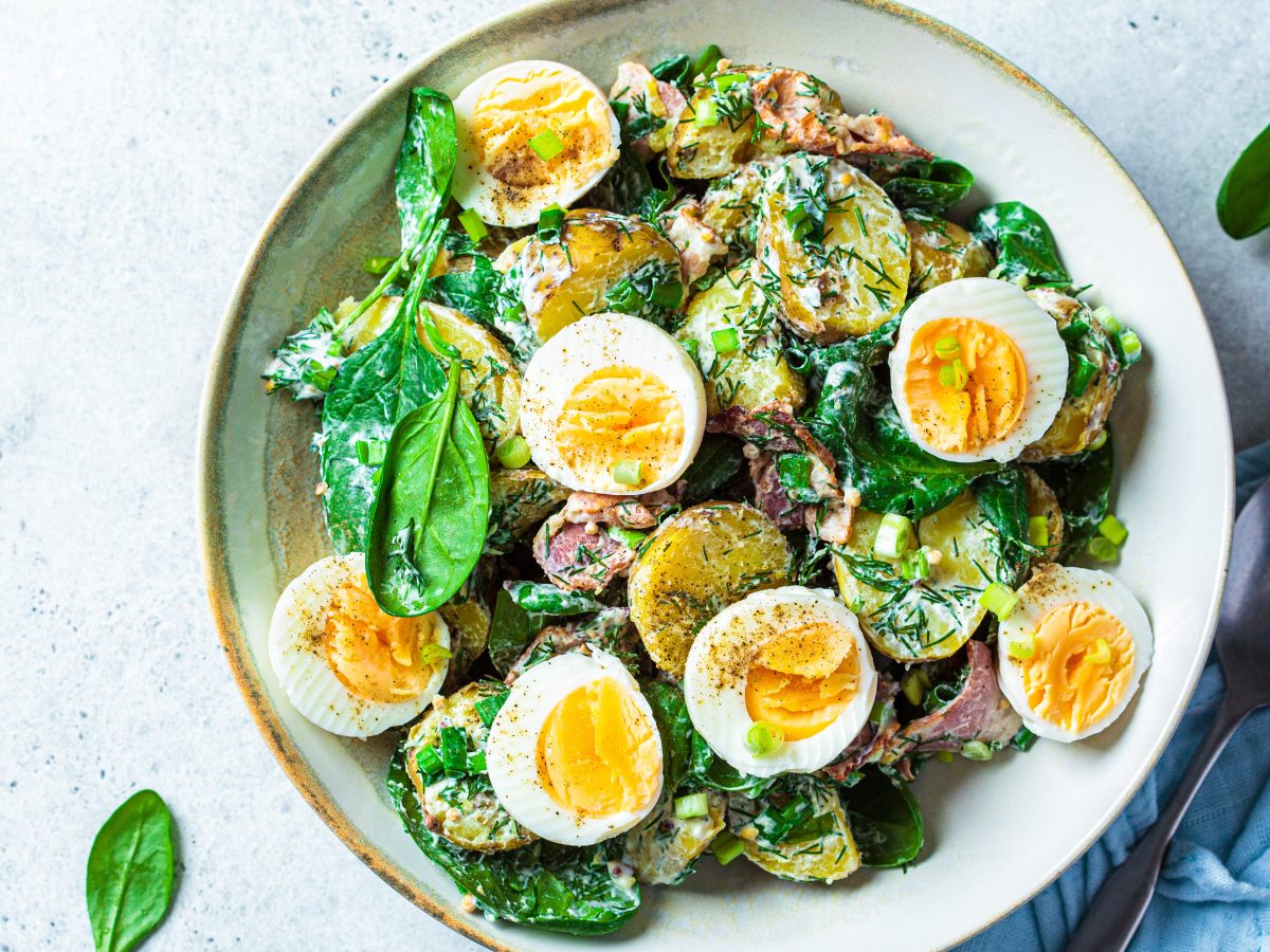 Eine weiße Schale mit dem Kartoffelsalat auf grau-blauem Untergrund und einer blauen Tischdecke mit silbernem Löffel unten links. Alles direkt von oben fotografiert.