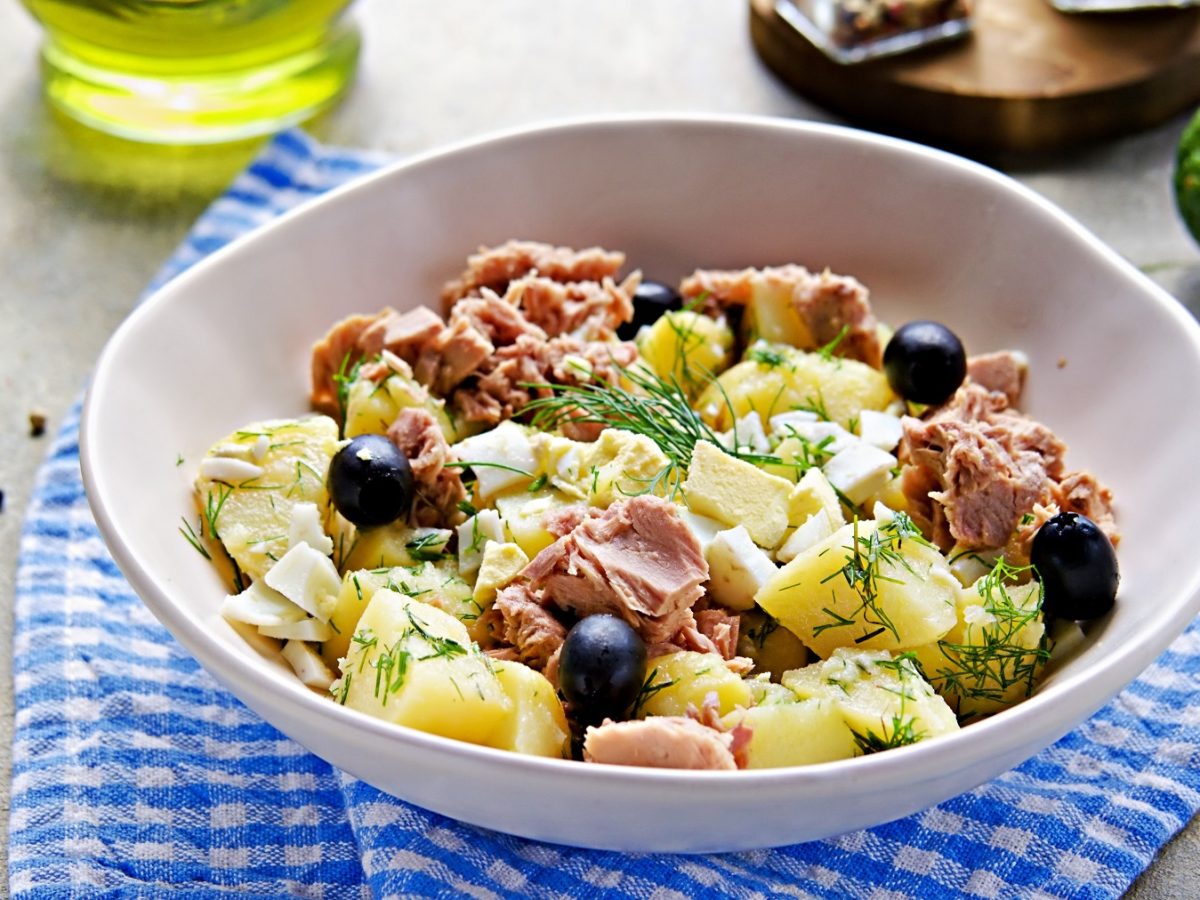 Eine weiße Schale mit dem Kartoffelsalat auf einem blau-weiß-gestreiftem Tuch und dunklem Holztisch. Alles von schräg oben fotografiert.