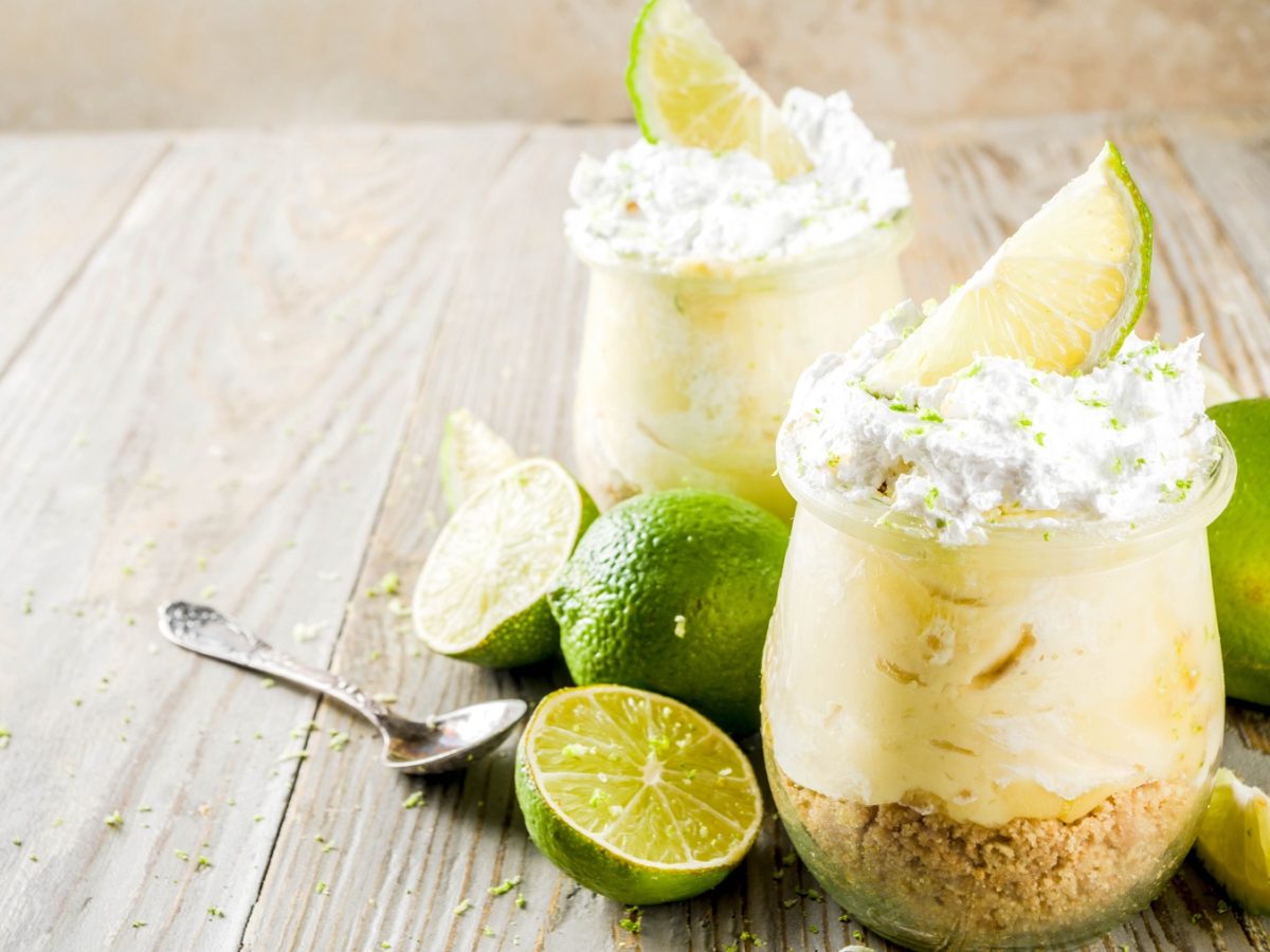 Zwei Gläser mit dem Key Lime Pie auf einem rustikalen Holztisch mit frischen Limetten und einem kleinen Löffel aus Metall. Alles von der Seite fotografiert.