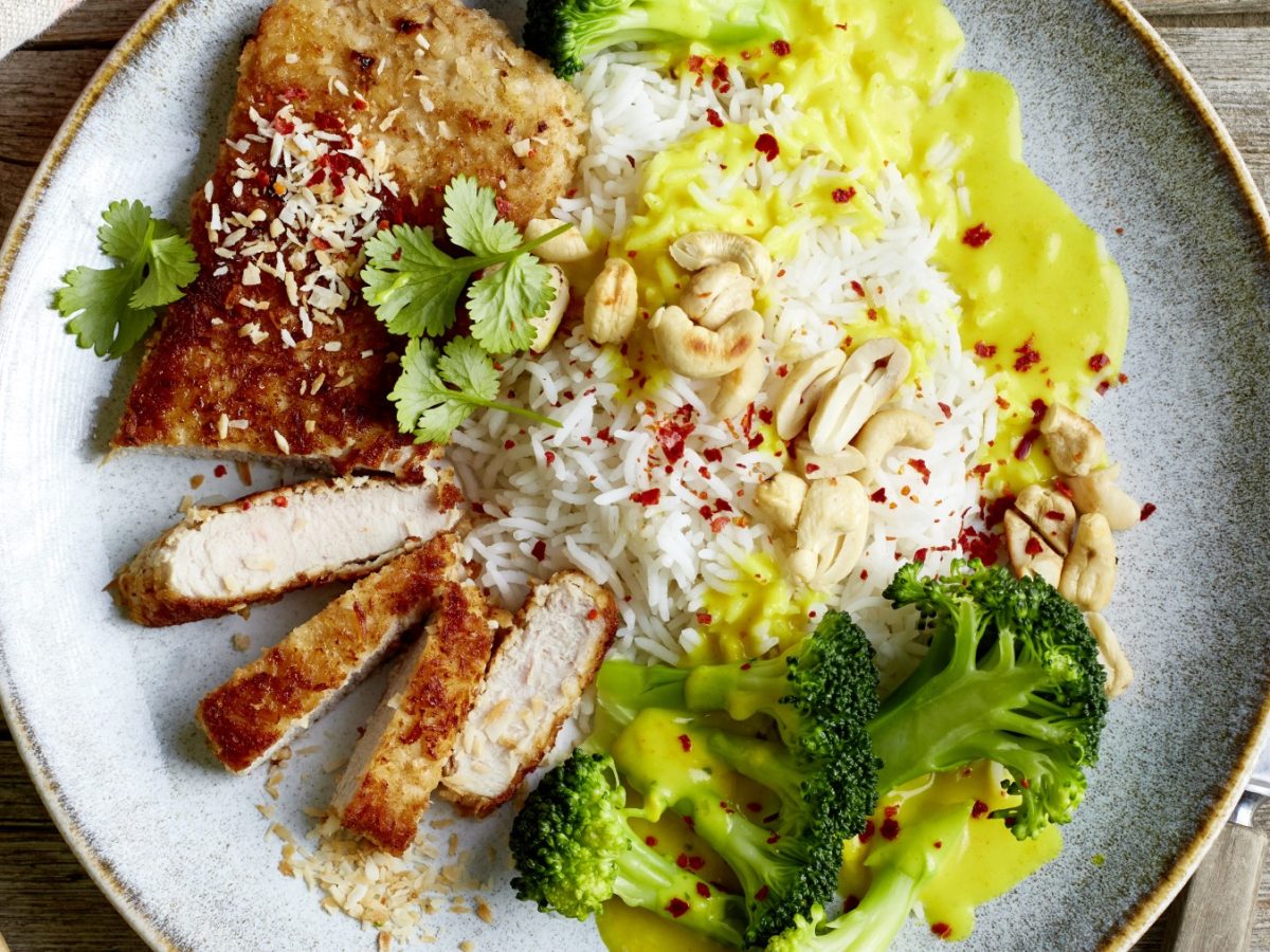 Kokosschnitzel mit Curryreis auf einem grauen Teller auf hölzernem Hintergrund. Rechts Besteck und links etwas Koriander.