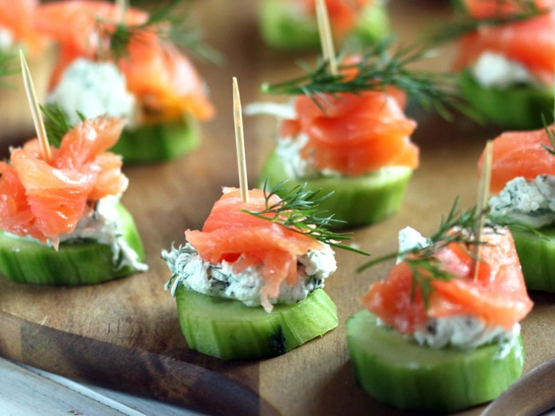 Mehrere Lachs-Häppchen auf einem dunklen Holzbrett von der Seite fotografiert.