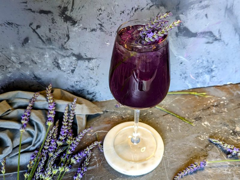 Ein Glas mit dem Lavendel Spritz auf grauem Untergrund mit frischen Lavendelzweigen und einem grauen Tuch. Alles von schräg oben fotografiert.