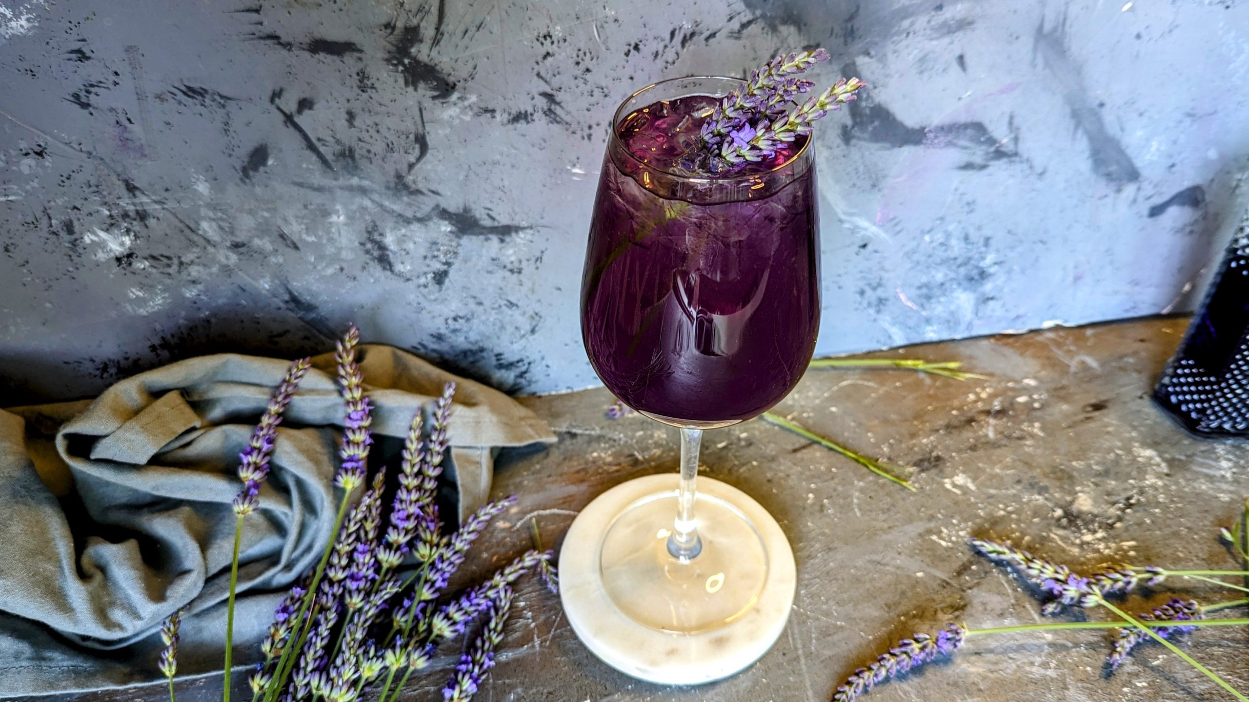 Ein Glas mit dem Lavendel Spritz auf grauem Untergrund mit frischen Lavendelzweigen und einem grauen Tuch. Alles von schräg oben fotografiert.