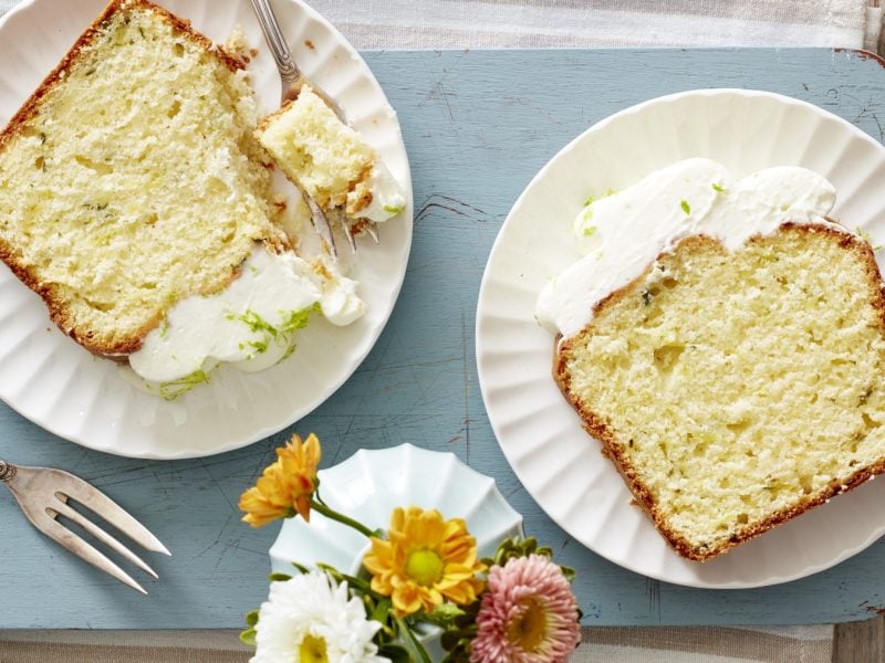 Limetten-Zucchini-Kuchen auf weißen Tellern auf blauem Hintergrund. Links unten eine Gabel und rechts davon Blumen.