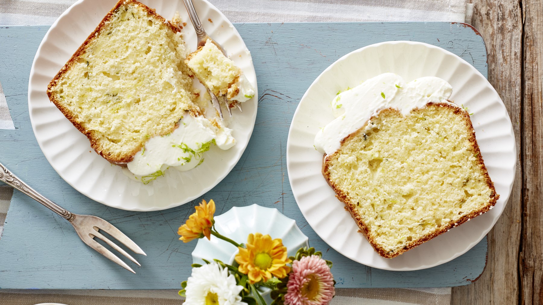 Limetten-Zucchini-Kuchen auf weißen Tellern auf blauem Hintergrund. Links unten eine Gabel und rechts davon Blumen.