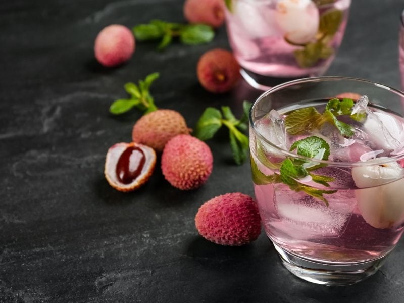 Drei Gläser mit Litschi Spritz auf dunkelm Untergrund, daneben frische Litschis in der Schale.