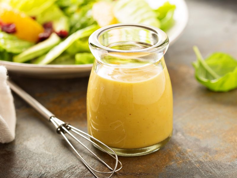 Ein Glas mit dem Apfel-Maracuja-Dressing auf eienm rustikalen Holztisch. Dahinter ein weißer Teller mit Blattsalat.