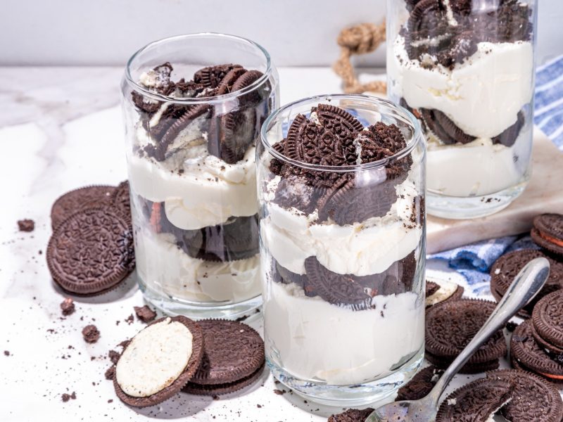 Drei Gläser mit dem Oreo-Trifle auf einem hellen Marmortisch mit blau-weiß-gestreifter Tischdecke.