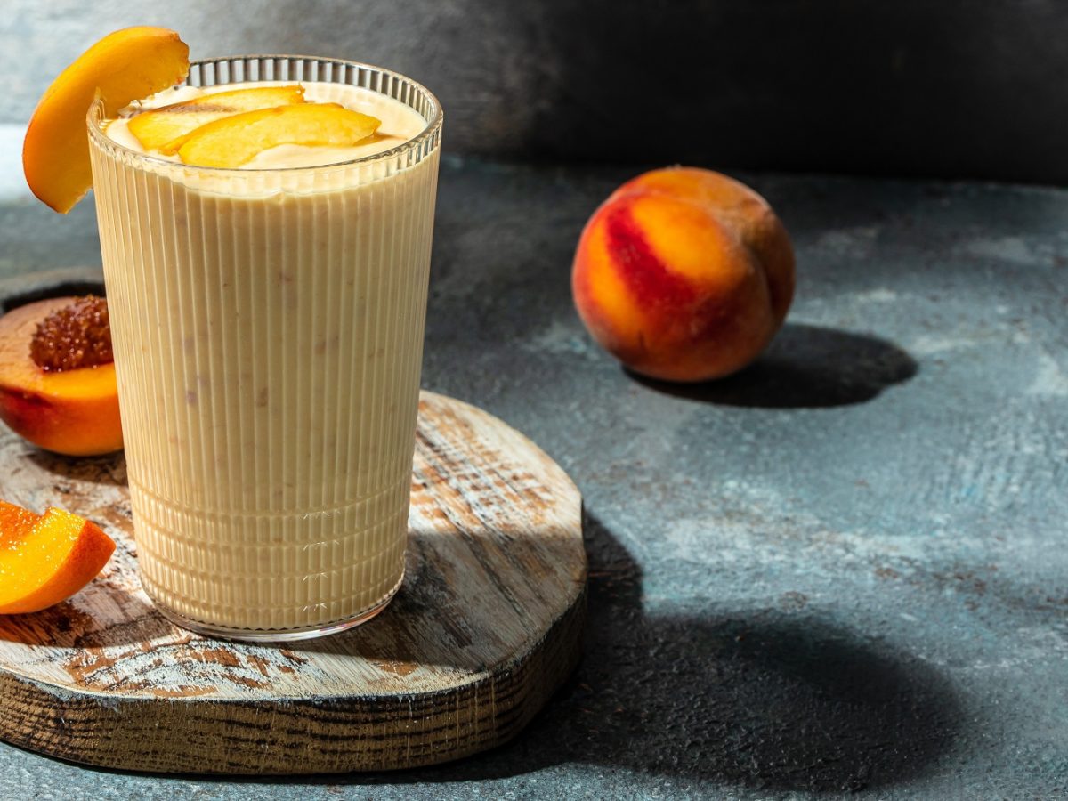 Ein Glas mit dem Shake auf einem Holzbrett und blau-grünlichem Marmorunter- und Hintergrund. Alles von der Seite fotografiert und drumherum frischer Pfirsich als Dekoration.