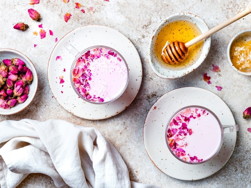 Zwei Tassen Pink Moon Milk mit Blütenblättern auf weißen Tellern, daneben Honig und Rosenknospen auf hellem Steinuntergrund.