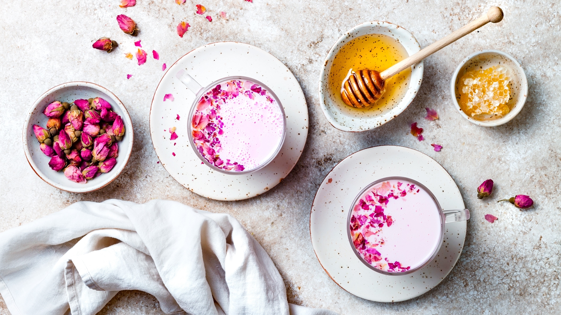 Zwei Tassen Pink Moon Milk mit Blütenblättern auf weißen Tellern, daneben Honig und Rosenknospen auf hellem Steinuntergrund.