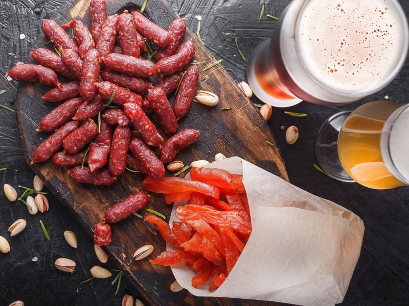 Eine Auswahl an roten Mini-Würstchen und geräuchertem Lachs auf Holzbrett mit Pistazien, begleitet von zwei Bieren.