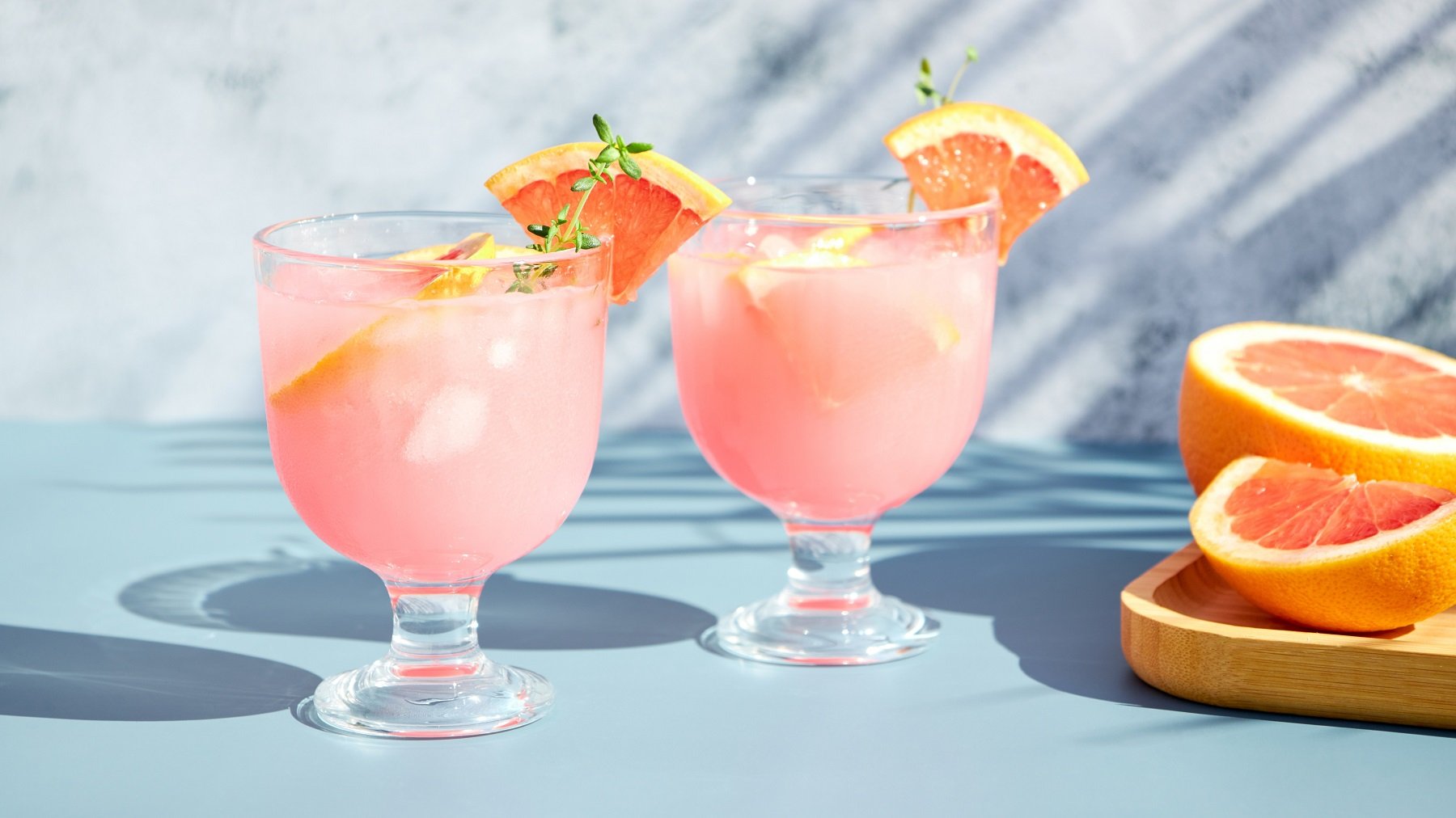 Zwei Gläser mit dem Drink auf hellblauem Unter- und Hintergrund. Rechts daneben angeschnittene Grapefruit auf einem Holzbrett.