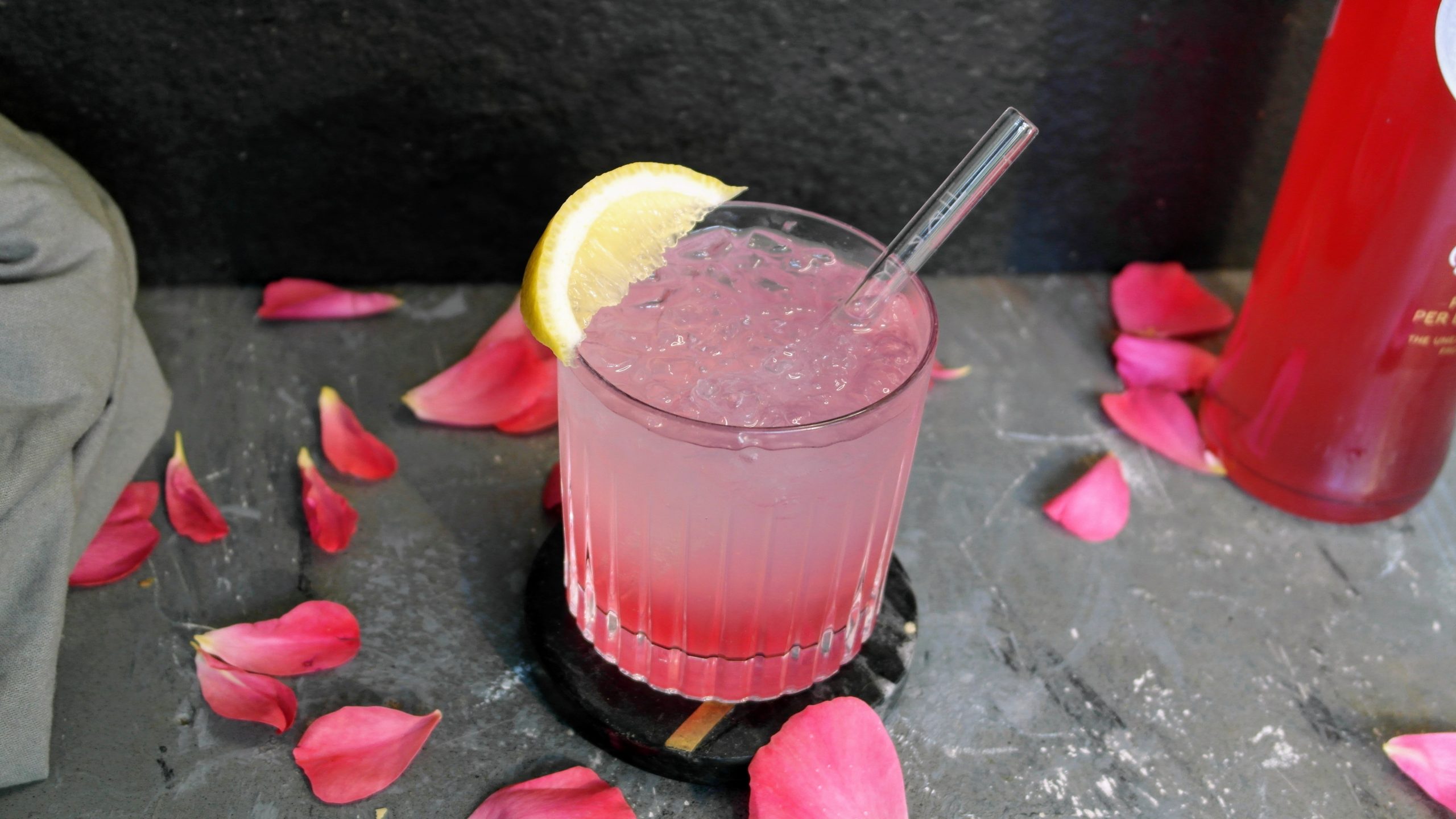 Ein Glas mit dem Sarti Lemon auf einem grauen Tisch mit pinken Blüten und einer Flasche Sarti. Alles vor schwarzem Hintergrund fotografiert.