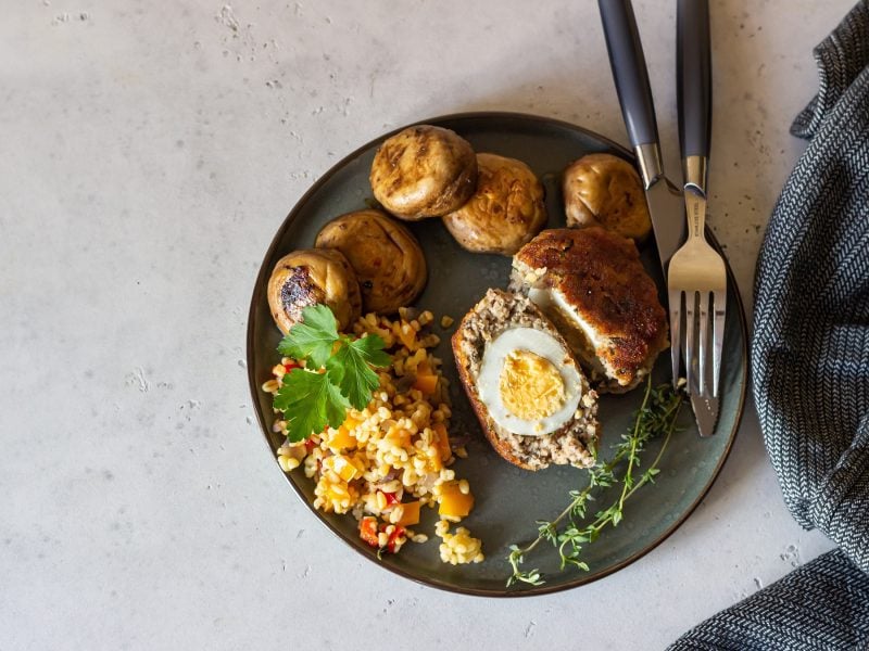 Ein Teller mit schottischen Eiern, gebackenen Champignons und buntem Couscous auf grauem Untergrund.