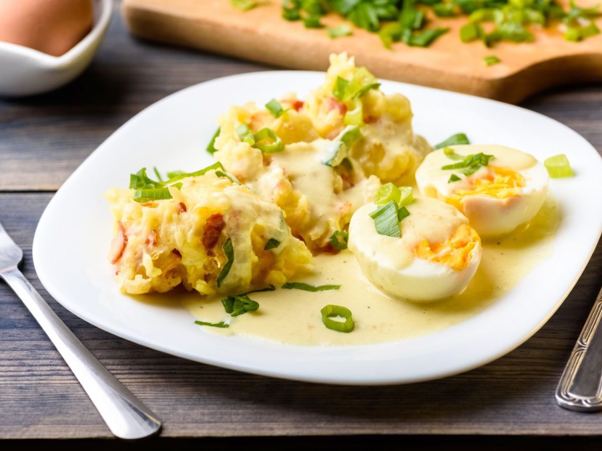 Kartoffelbrei mit halbierten Eiern in Send-Sauce und Frühlingszwiebeln auf weißem Teller, daneben Messer und Gabel.