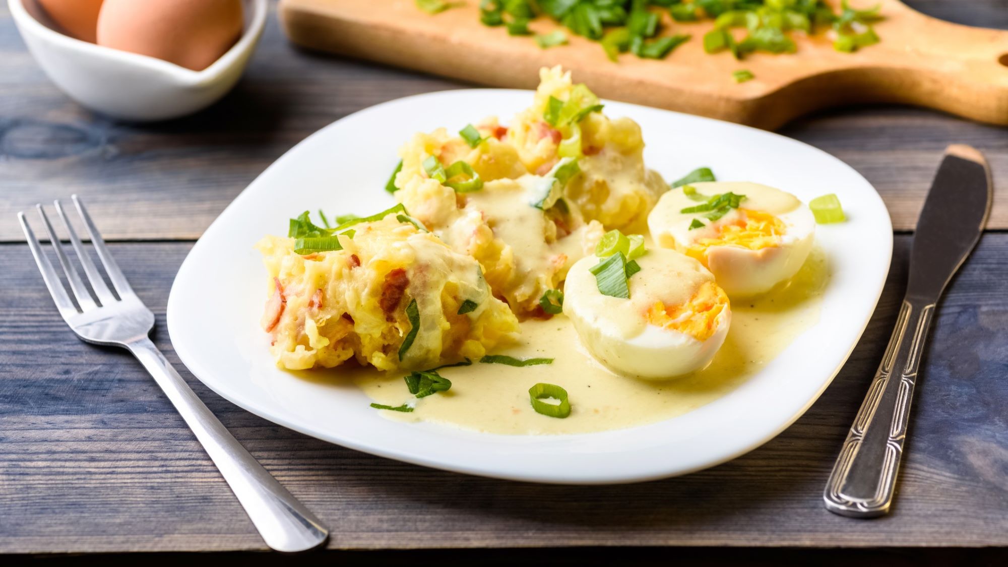 Kartoffelbrei mit halbierten Eiern in Send-Sauce und Frühlingszwiebeln auf weißem Teller, daneben Messer und Gabel.