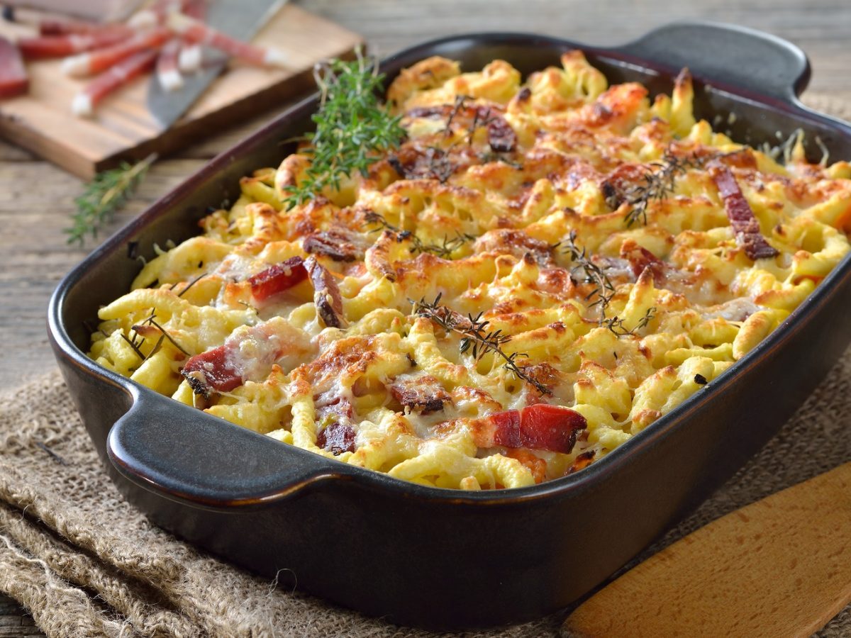 Eine schwarze Auflaufform mit dme Spätzle-Gratin auf einem rustikalen Holztisch. Im Hintergrund unscharf ganzer Speck zu erkennen. Alles von leicht schräg oben fotografiert.