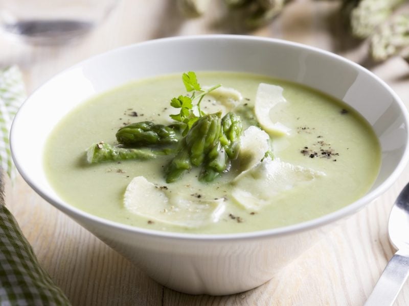 Spargelcremesuppe mit Parmesan in weißer Schüssel vor hellem Stoff-Hintergrund, daneben ein Löffel.