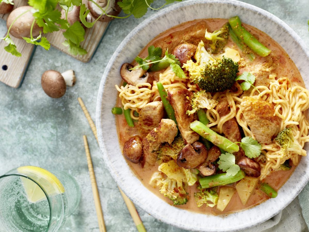 Thai-Curry mit Nudeln in einer weißen Schüssel angerichtet. Der Hintergrund ist grau und darauf sind Koriander und Champignons verteilt. Links unten auf dem Foto ein Glas gefüllt mit Wasser.