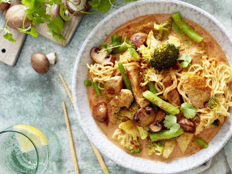 Thai-Curry mit Nudeln in einer weißen Schüssel angerichtet. Der Hintergrund ist grau und darauf sind Koriander und Champignons verteilt. Links unten auf dem Foto ein Glas gefüllt mit Wasser.