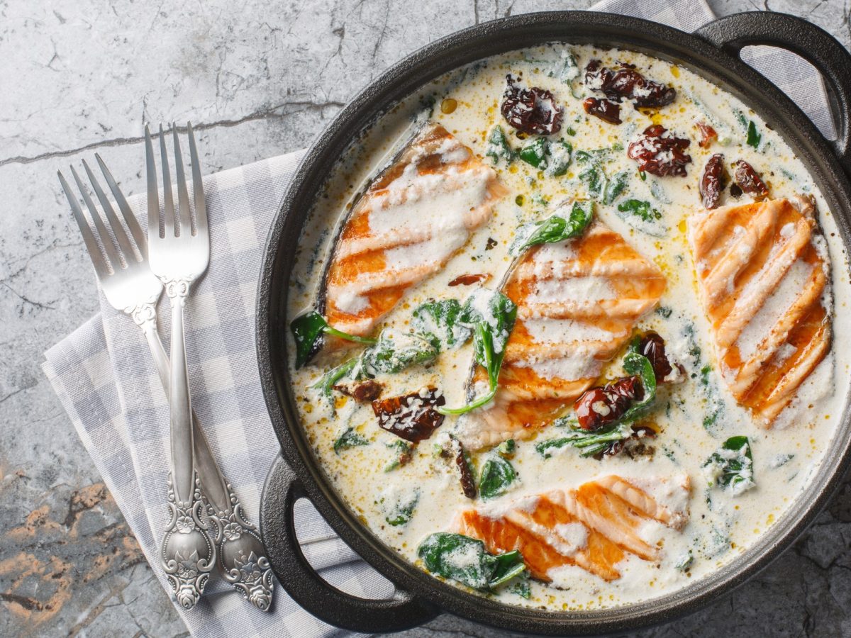 Eine Pfanne mit gegrilltem Lachs in cremiger Sauce mit Spinat und getrockneten Tomaten, auf grauem Marmorhintergrund, zwei silberne Gabeln daneben.