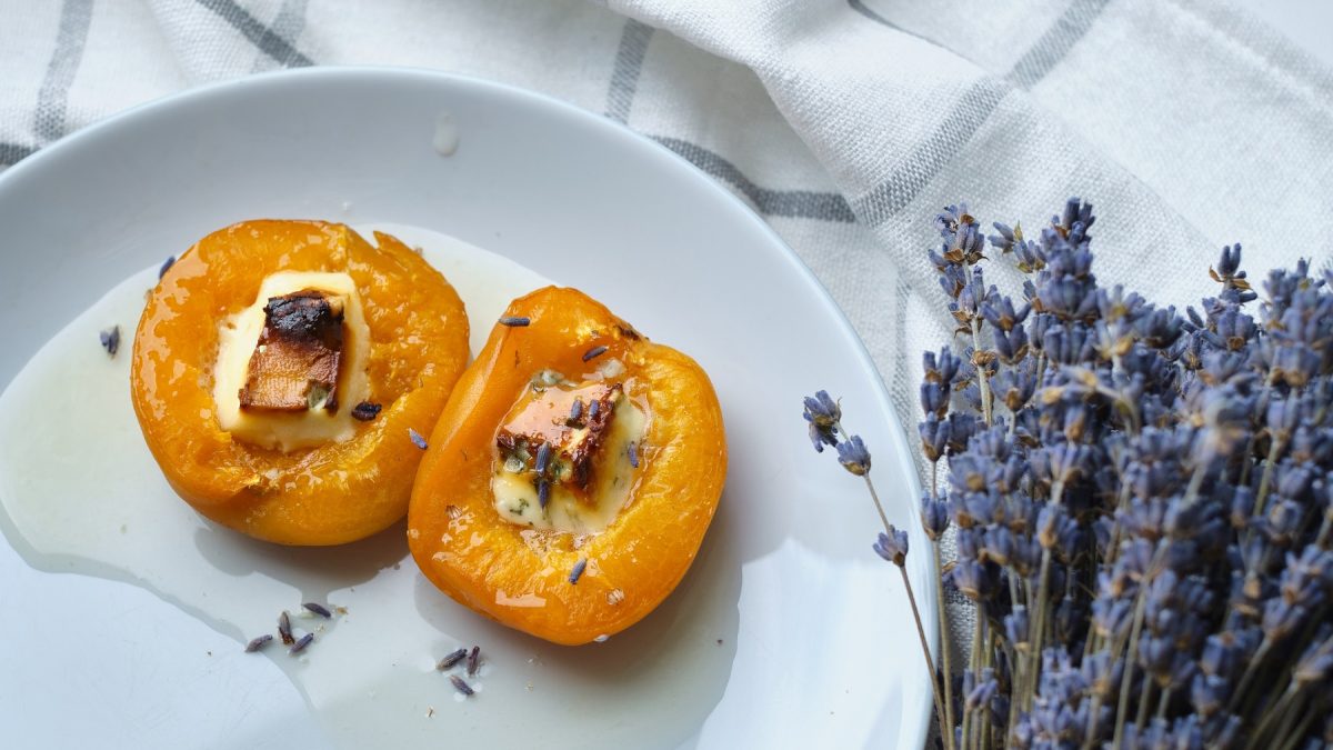 Zwei überbackene Aprikosenhälften mit Käse, Honig und Lavendelblüten bestreut auf einem weißen Teller und eine blau-weiß-karierten Tischdecke. Rechts daneben ein Strauß aus Lavendel