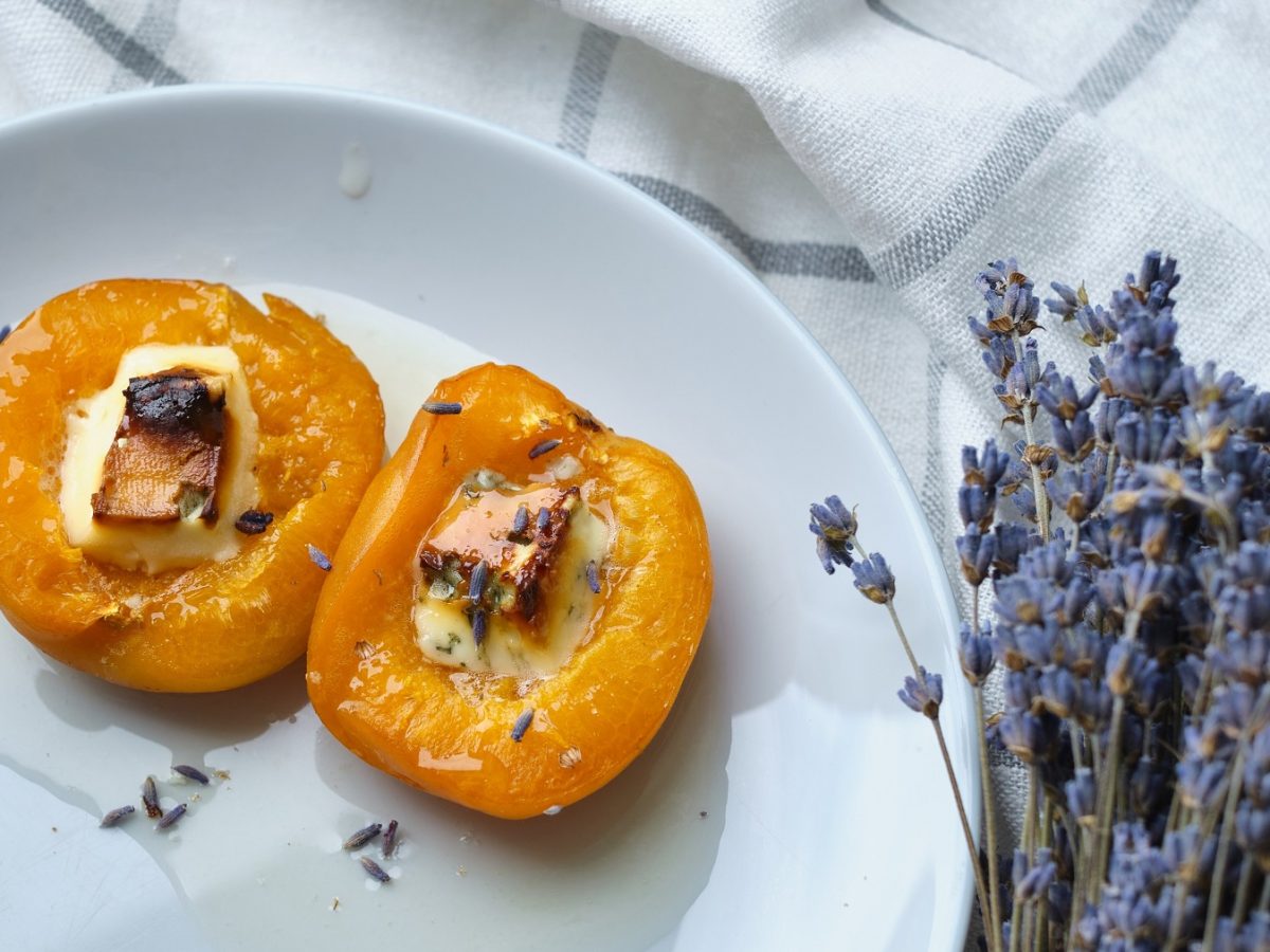 Zwei überbackene Aprikosenhälften mit Käse, Honig und Lavendelblüten bestreut auf einem weißen Teller und eine blau-weiß-karierten Tischdecke. Rechts daneben ein Strauß aus Lavendel