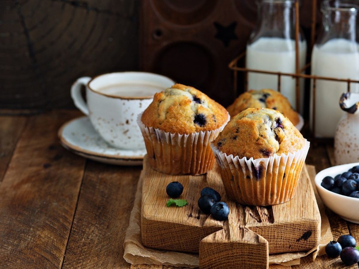 Zwei vegane Heidelbeer-Muffins auf einem Holzbrett, daneben ein Becher Kaffee, Blaubeeren in einer Schale und Milchflaschen im Hintergrund.