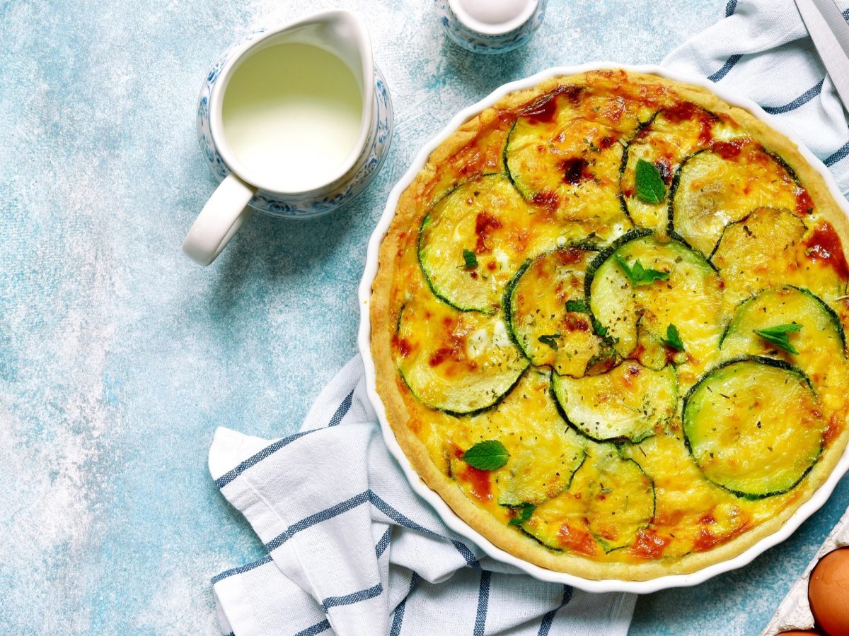 Draufsicht: Eine runde, weiß Form mit einer Zucchini-Quiche mit Käse auf hellblauem Untergrund. Ebenfalls zu sehen sind frische Eier, ein Küchenmesser, ein Kännchen mit Sahne und ein Küchentuch.