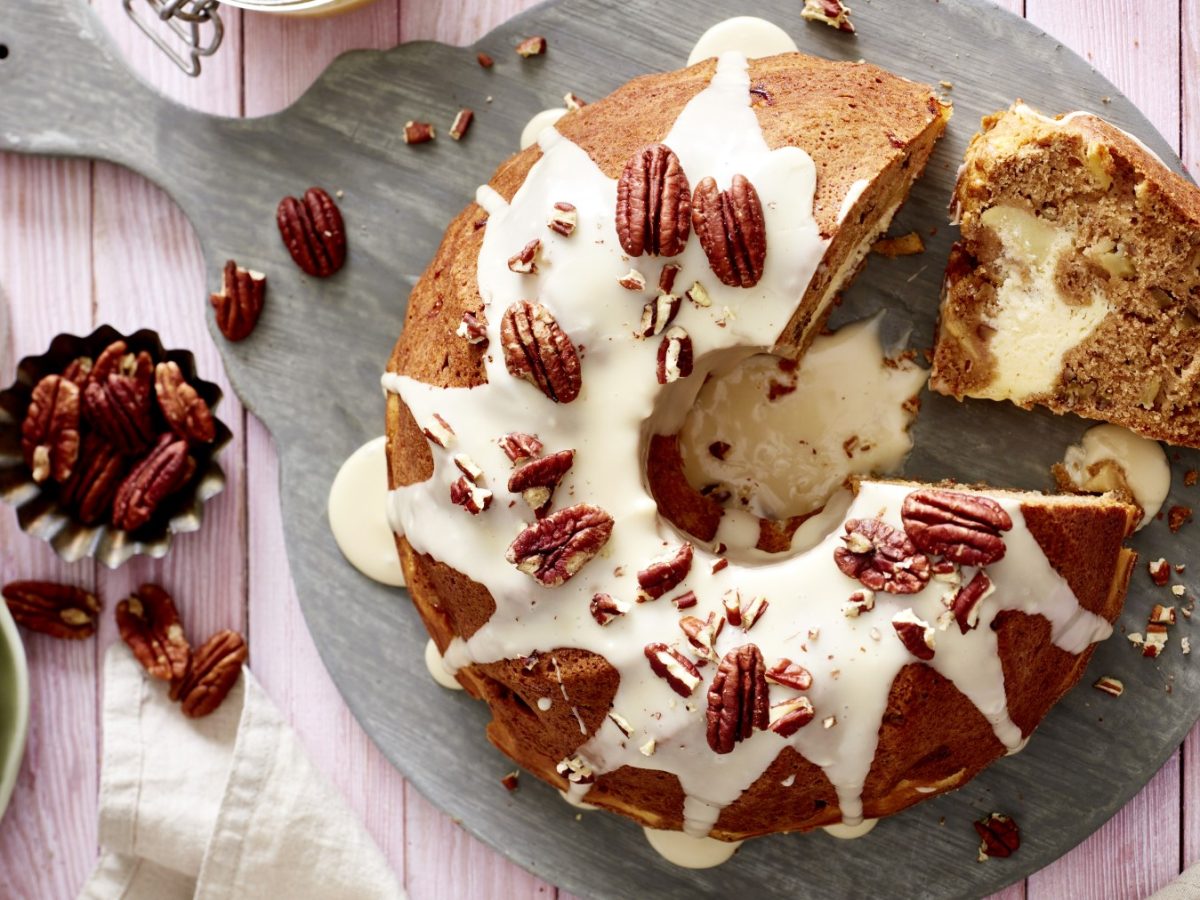 Apfel-Gugel mit Cheesecake Füllung auf einem grauen Servierteller auf rosarotem Hintergrund. Links davon Blumen, Geschirr und Pekannüsse.