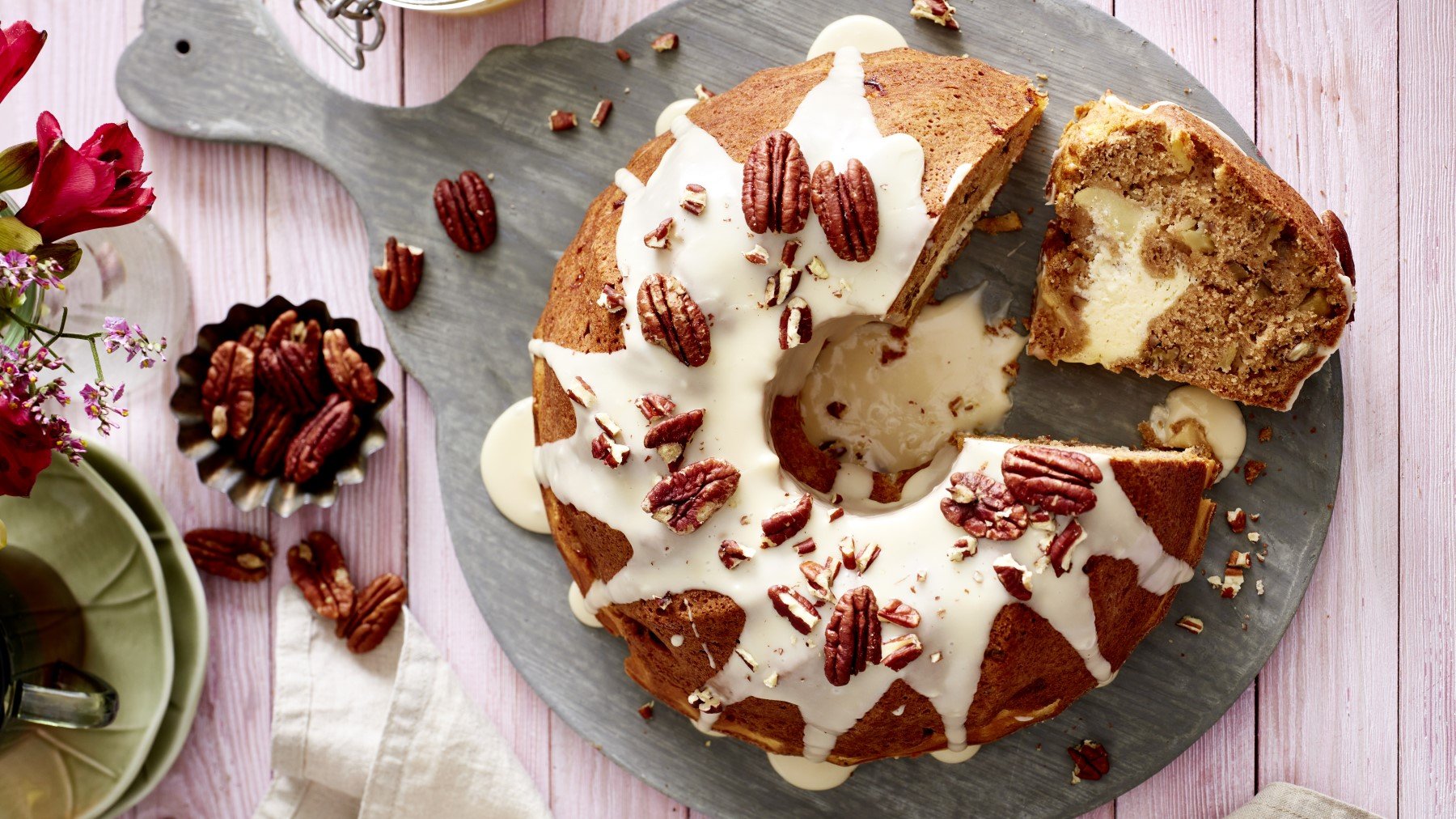 Apfel-Gugel mit Cheesecake Füllung auf einem grauen Servierteller auf rosarotem Hintergrund. Links davon Blumen, Geschirr und Pekannüsse.