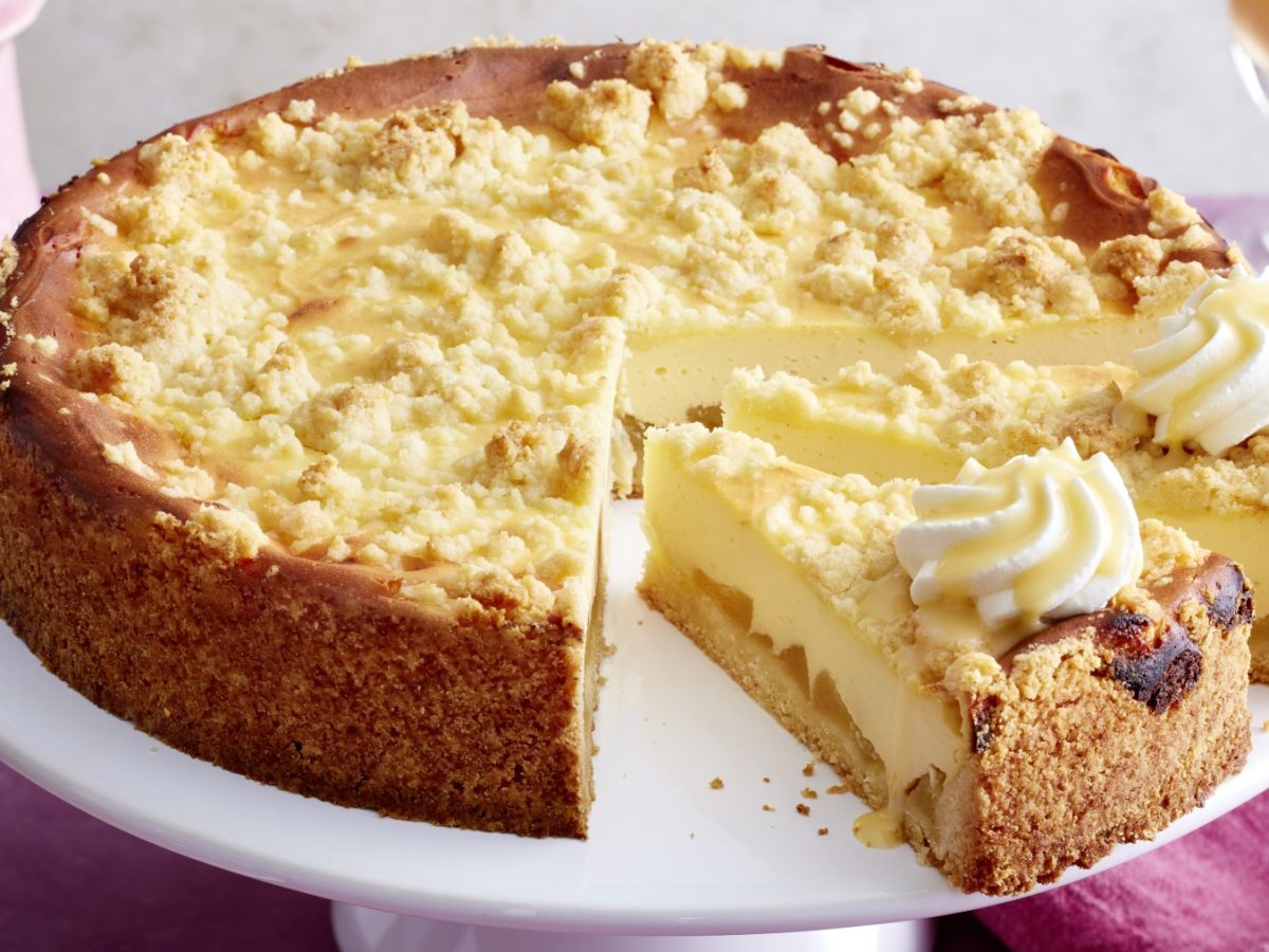 Angeschnittener Apfel-Käsekuchen mit Streuseln und Sahne auf Servierplatte.