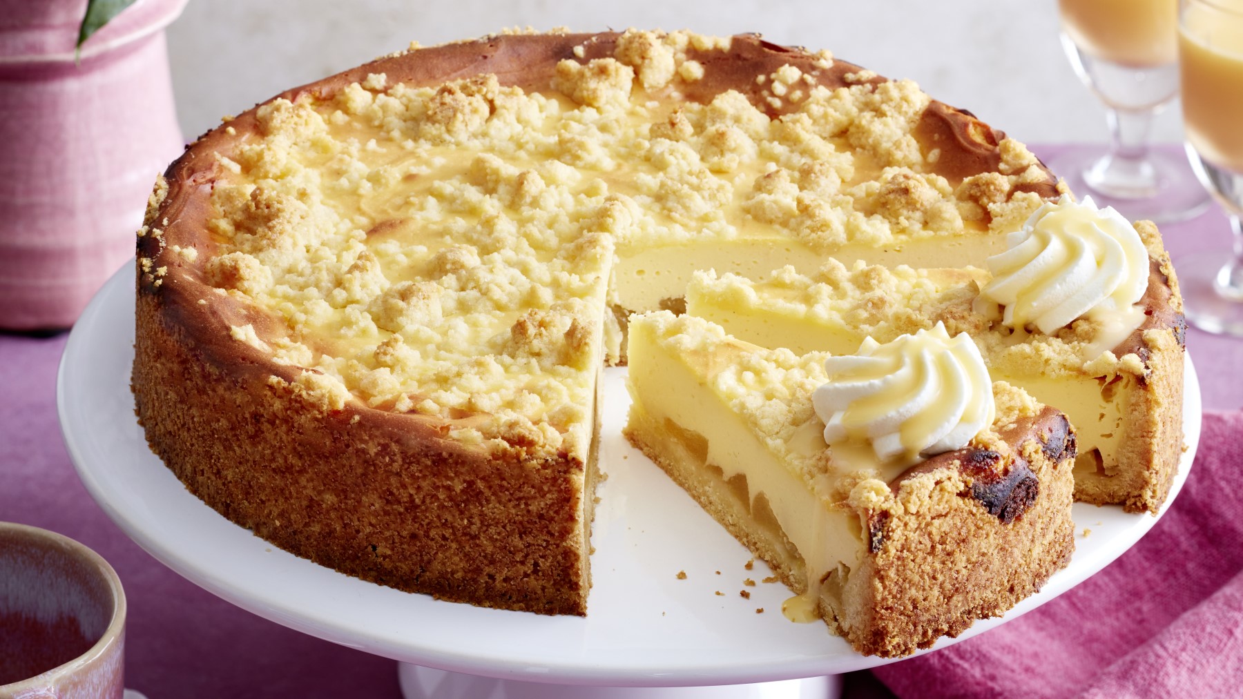 Angeschnittener Apfel-Käsekuchen mit Streuseln und Sahne auf Servierplatte.