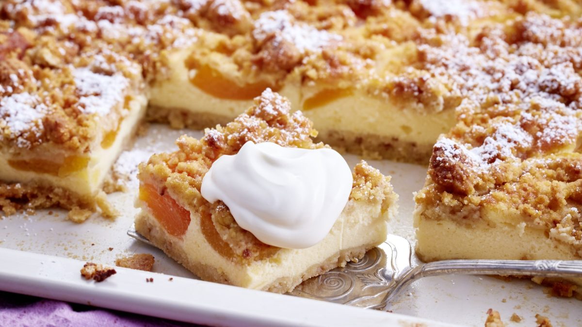 Angeschnittener Aprikosen-Schmandkuchen mit Schlagsahne auf weißem Blech.