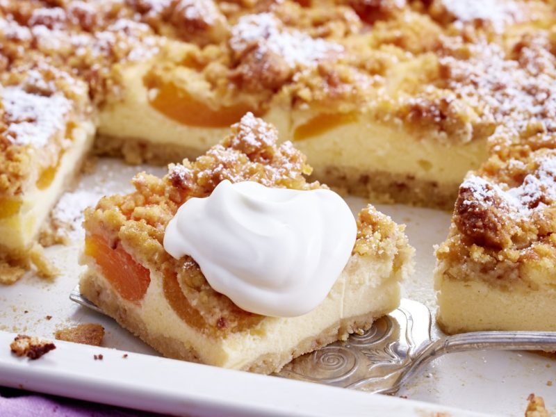 Angeschnittener Aprikosen-Schmandkuchen mit Schlagsahne auf weißem Blech.