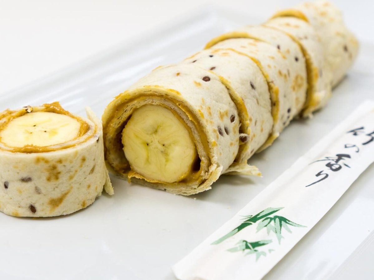 Das geschnittene Bananen-Sushi in naher Ansicht auf einem weißen, rechteckigen Teller schräg von der Seite, mit Essstäbchen und weißem Hintergrund fotografiert.