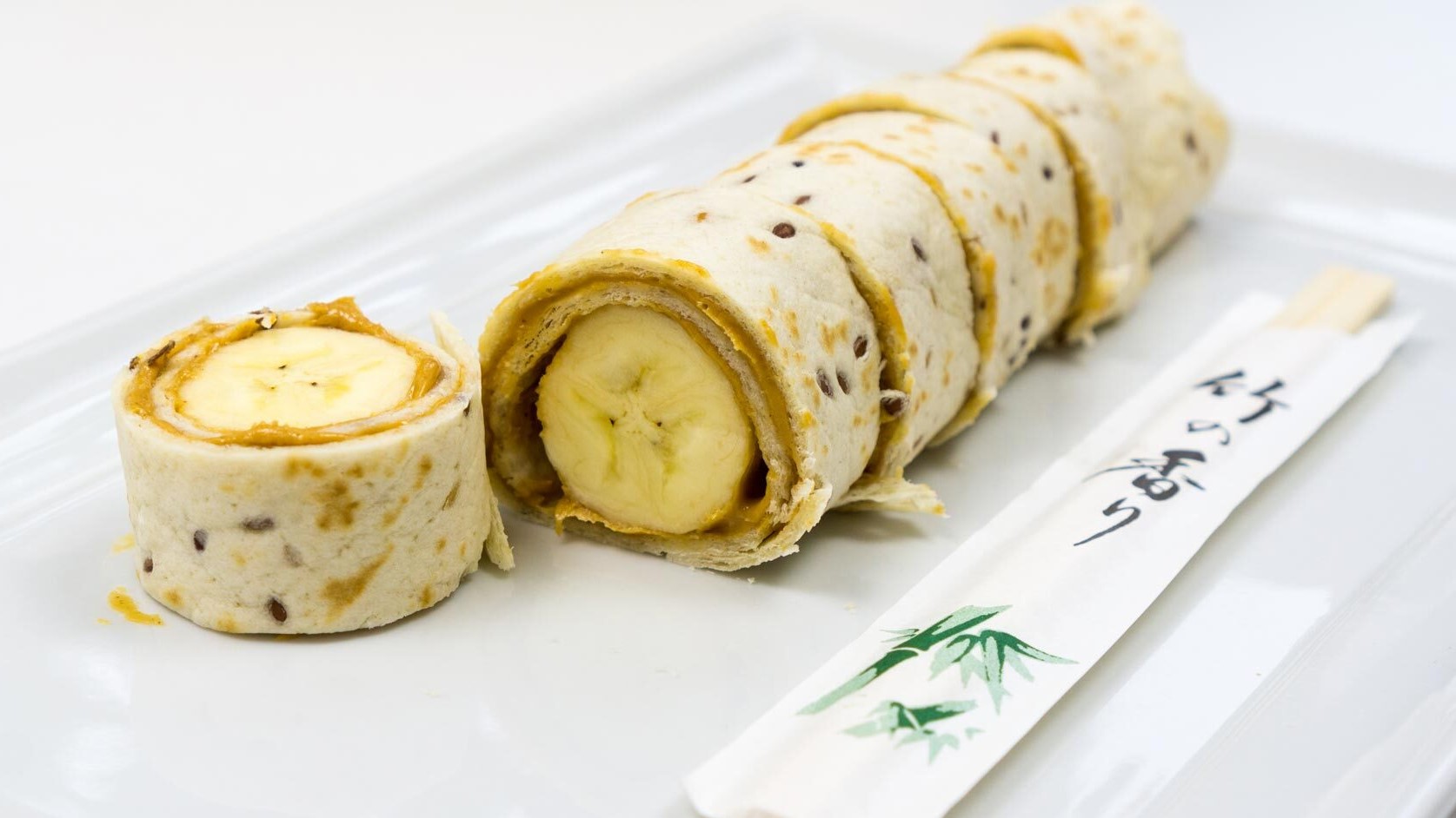 Das geschnittene Bananen-Sushi in naher Ansicht auf einem weißen, rechteckigen Teller schräg von der Seite, mit Essstäbchen und weißem Hintergrund fotografiert.