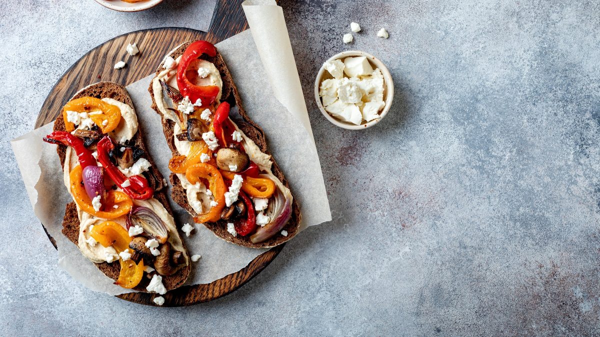 Zwei Brotscheiben mit Hummus, gegrillten Paprikaschoten, Zwiebeln und Fetastückchen auf einem Holzbrett.