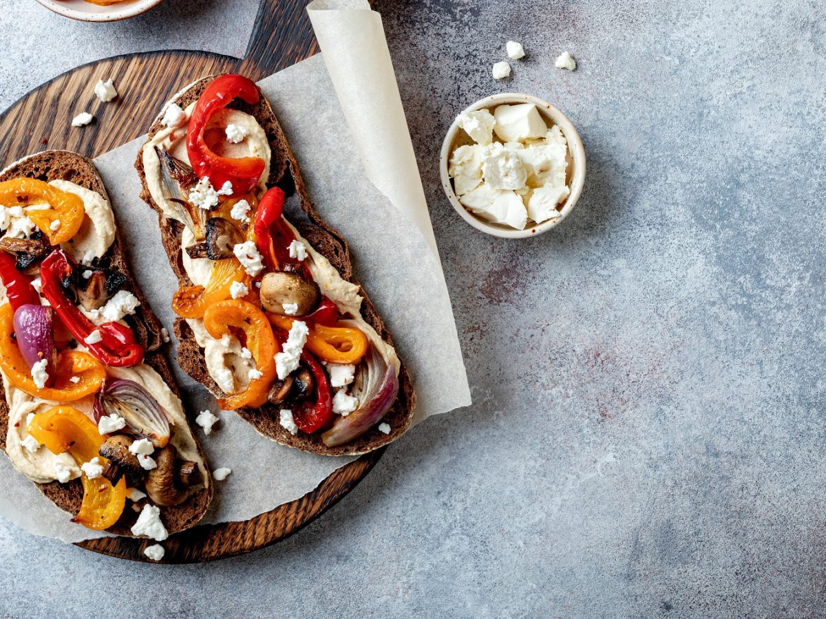 Zwei Brotscheiben mit Hummus, gegrillten Paprikaschoten, Zwiebeln und Fetastückchen auf einem Holzbrett.