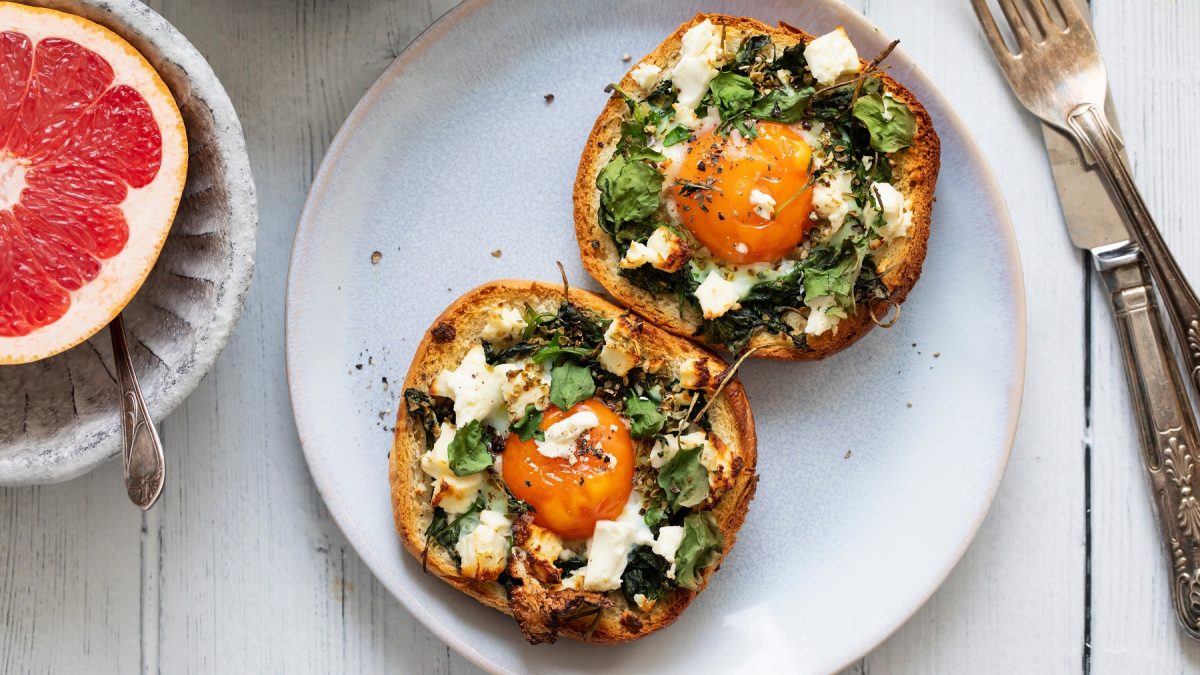 Zwei belegte Brothälften mit Spinat, Feta und Spiegelei auf einem hellblauen Teller, daneben eine halbe Grapefruit.