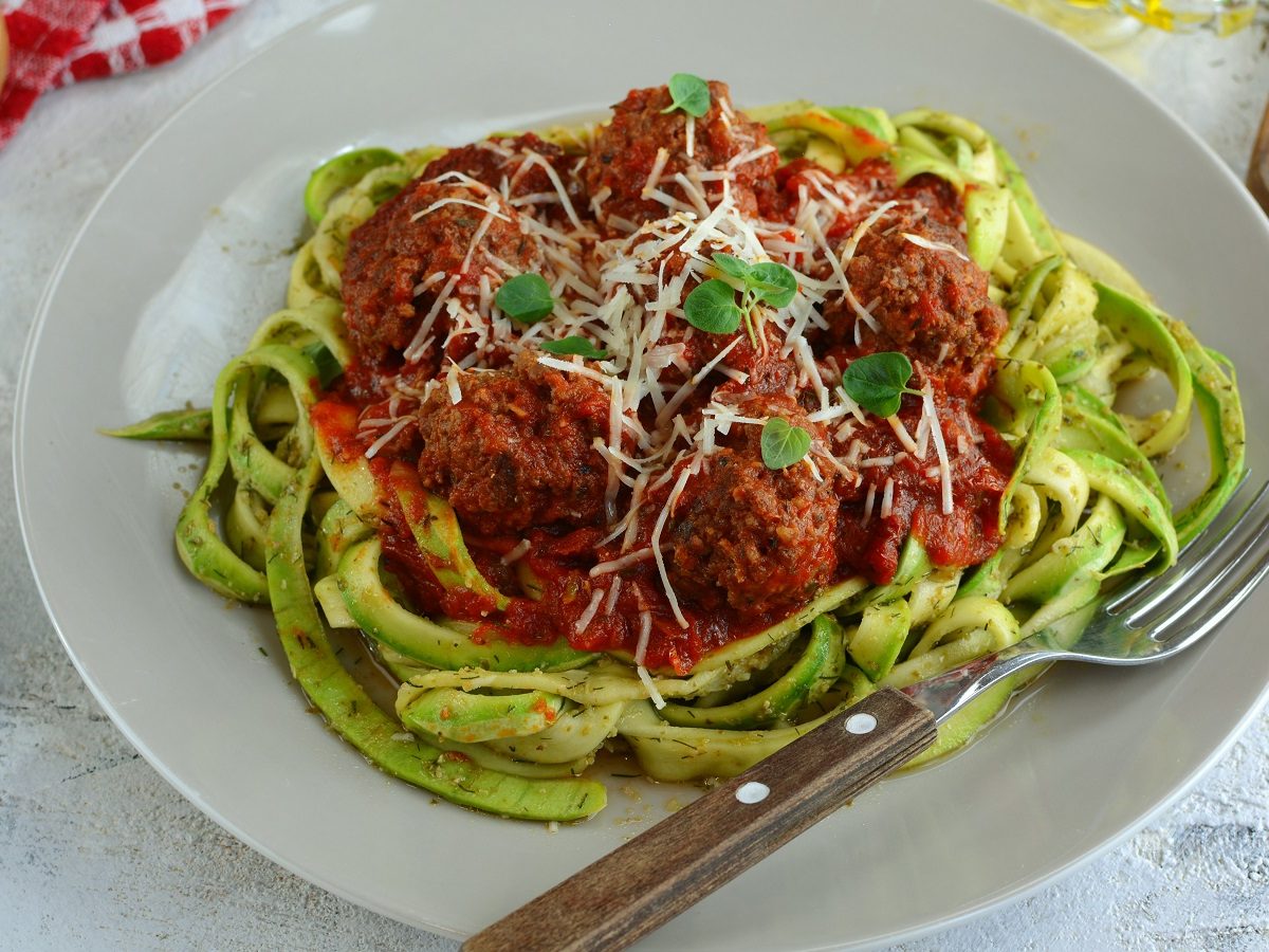 Ein heller Teller mit den grünen Nudeln und der Tomatensauce, mit Hackbällchen auf hellem Untergrund, mit allerhand mediterraner Deko drumherum. Alles von schräg oben fotografiert.