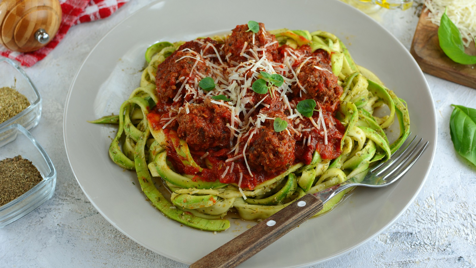 Ein heller Teller mit den grünen Nudeln und der Tomatensauce, mit Hackbällchen auf hellem Untergrund, mit allerhand mediterraner Deko drumherum. Alles von schräg oben fotografiert.