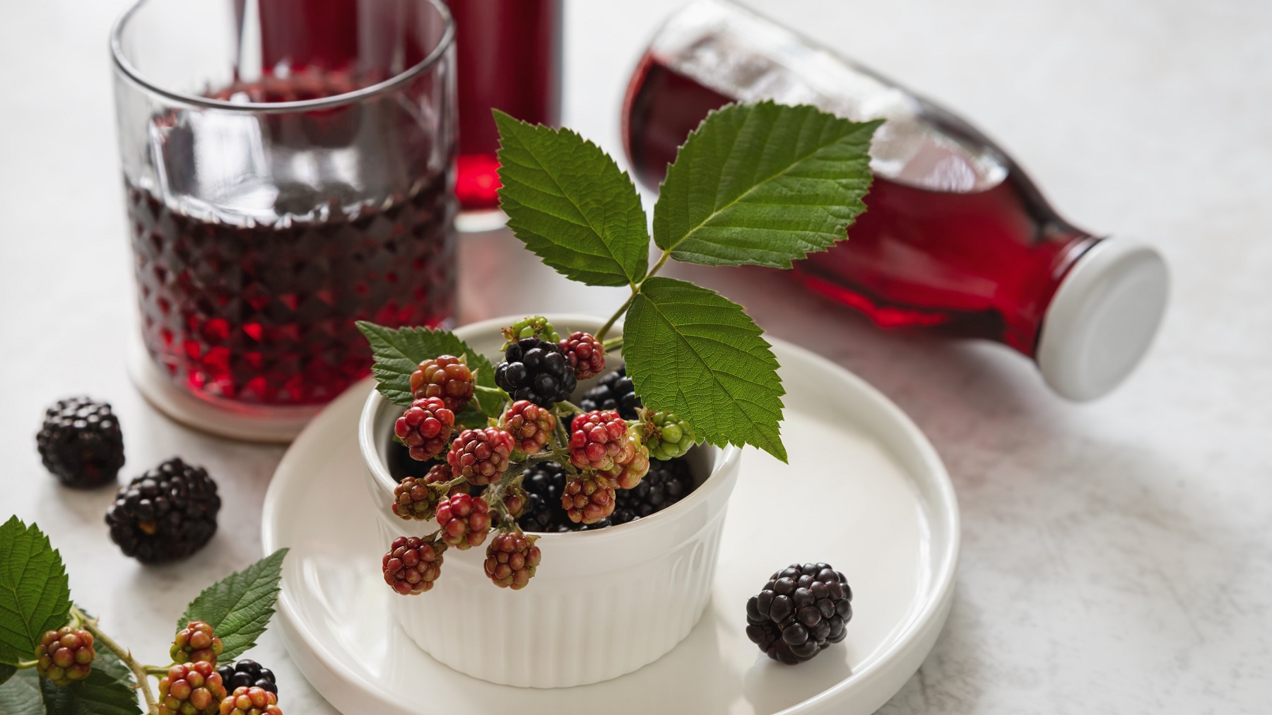 Ein weißer Teller mit frischen Brombeeren und im Hintergrund der Likör in Flaschen und Gläsern abgefüllt.