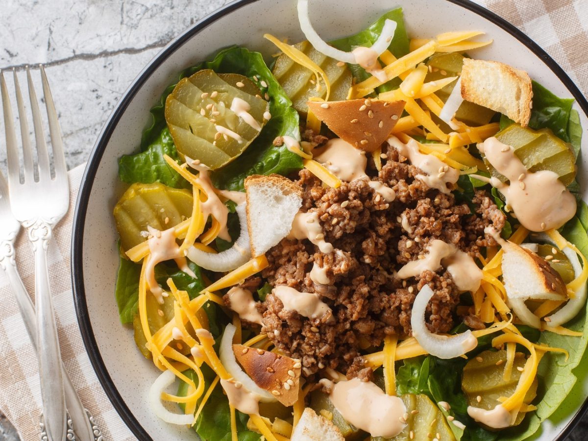 Eine weiße Schüssel mit schwarzem Rand und dem Cheeseburger-Salat auf einem grauen Tisch mit braun-weiß karierter Tischdecke. Alles direkt von fotografiert.