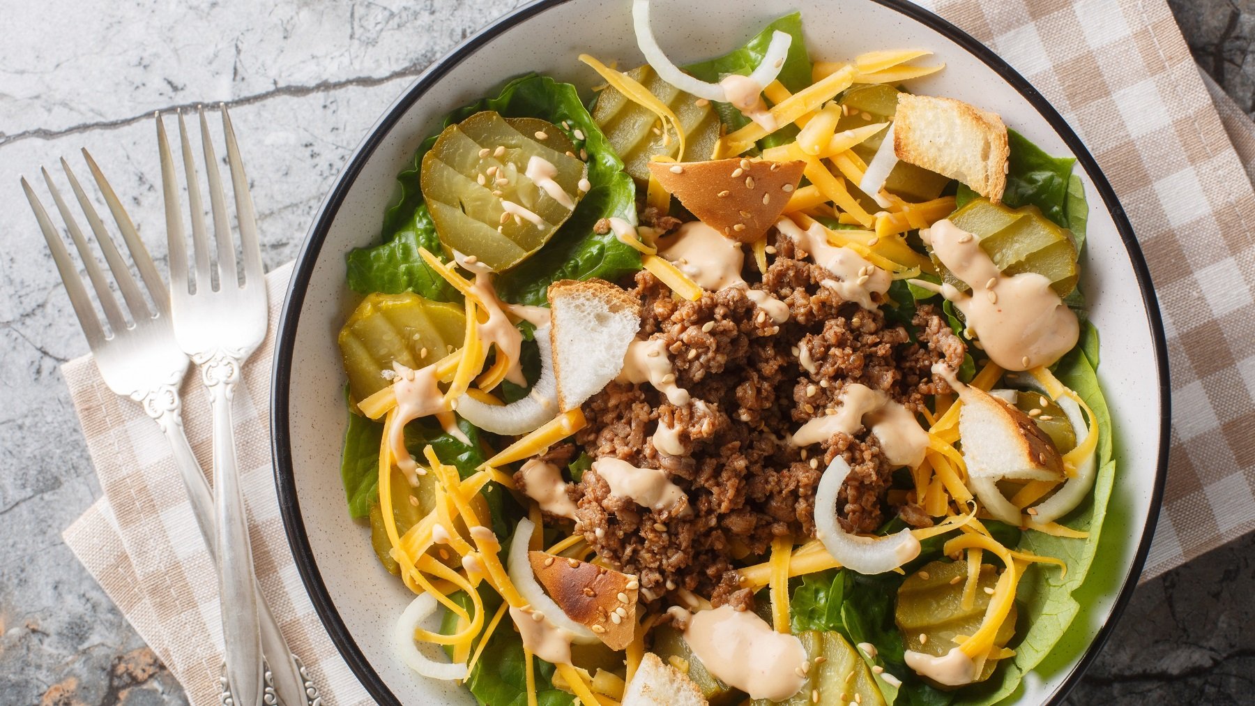 Eine weiße Schüssel mit schwarzem Rand und dem Cheeseburger-Salat auf einem grauen Tisch mit braun-weiß karierter Tischdecke. Alles direkt von fotografiert.