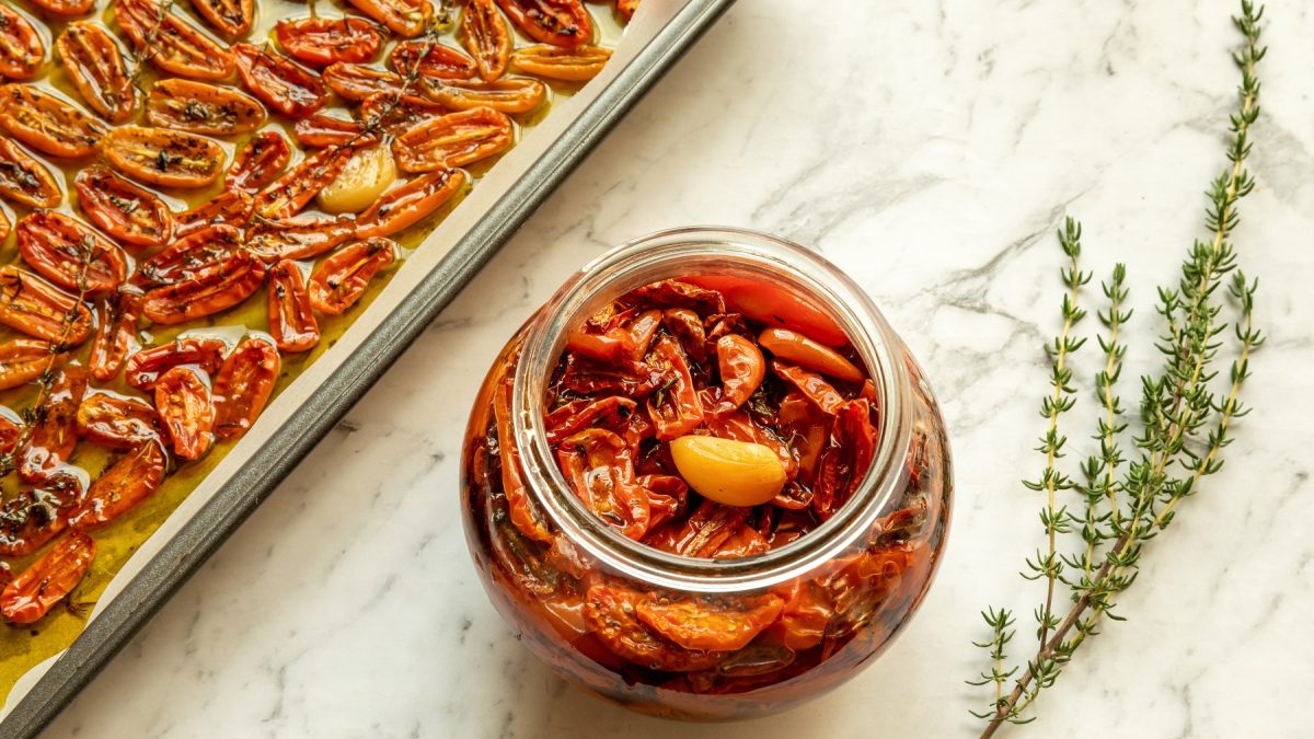 Glas mit confierten Tomaten und Knoblauch neben einem Backblech mit Tomaten und zwei Thymianzweigen auf hellem Marmor.