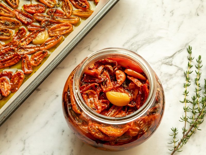 Glas mit confierten Tomaten und Knoblauch neben einem Backblech mit Tomaten und zwei Thymianzweigen auf hellem Marmor.