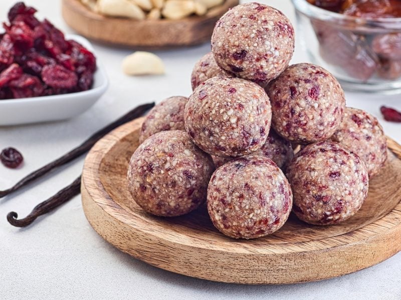 Cranberry-Energyballs auf einem Holzteller. Im Hintergrund die Zutaten für die Bällchen.