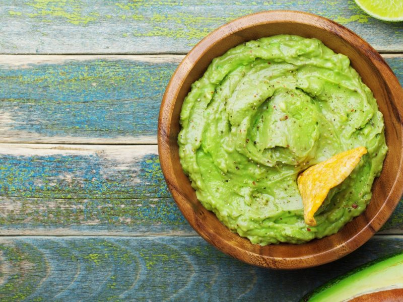 Draufsicht: Eine blauer-mediterraner Untergrund aus Holz. Auf dem steht eine Schüssel mit einem Avocado-Dip mit Limette. Daneben liegen eine halbe Avocado und zwei Limetten-Schnitzer,